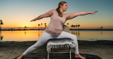 A woman is seen stretching outdoors with the help of a chair as the sun rises over a calm body of water behind her.