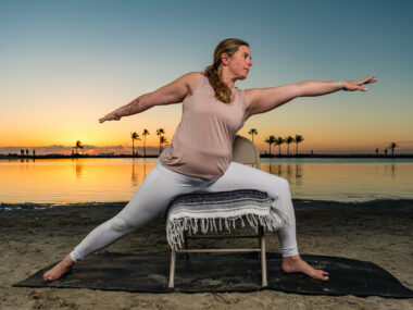 A woman is seen stretching outdoors with the help of a chair as the sun rises over a calm body of water behind her.