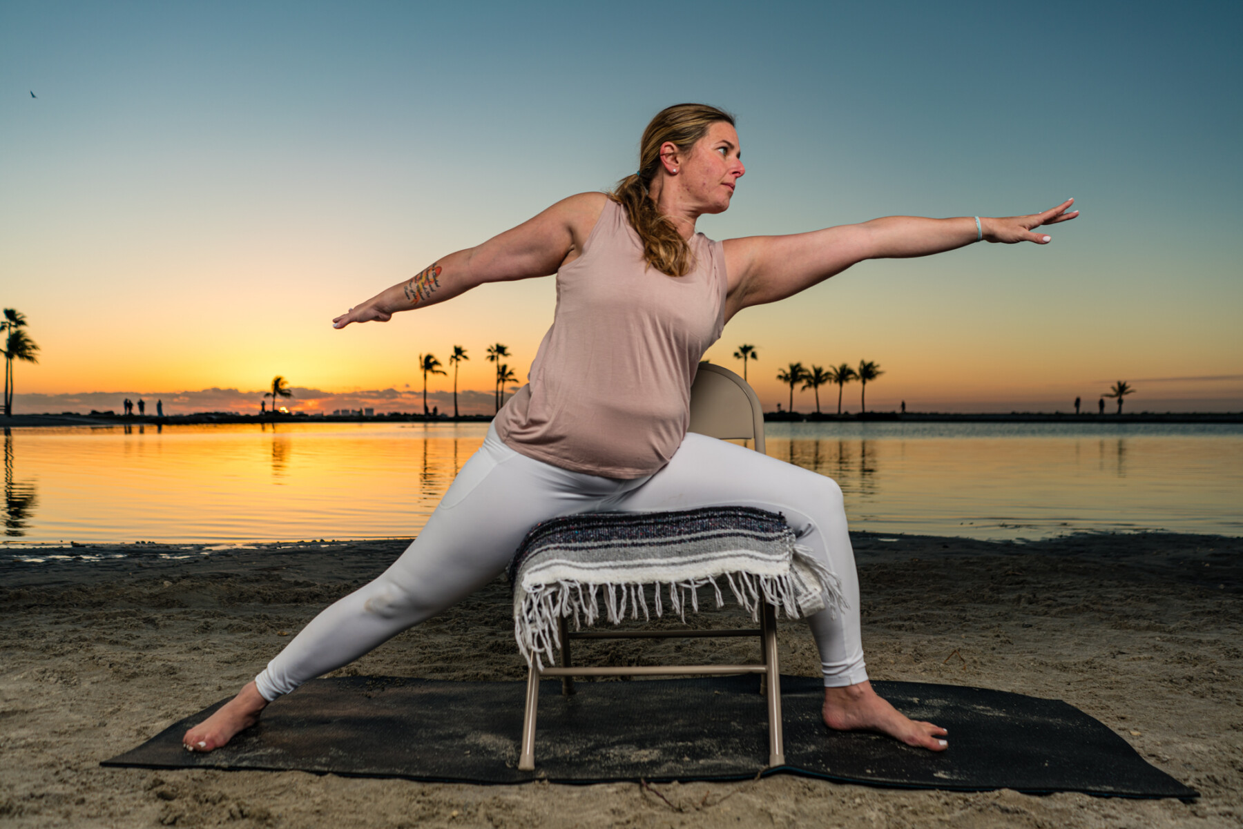 A woman is seen stretching outdoors with the help of a chair as the sun rises over a calm body of water behind her.