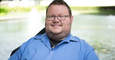 A pretty tight horizontal profile pic of a man in a blue dress shirt, short-cropped hair, and glasses, smiling broadly.