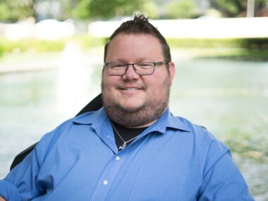 A pretty tight horizontal profile pic of a man in a blue dress shirt, short-cropped hair, and glasses, smiling broadly.