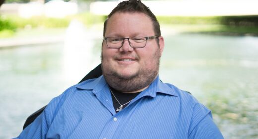A pretty tight horizontal profile pic of a man in a blue dress shirt, short-cropped hair, and glasses, smiling broadly.
