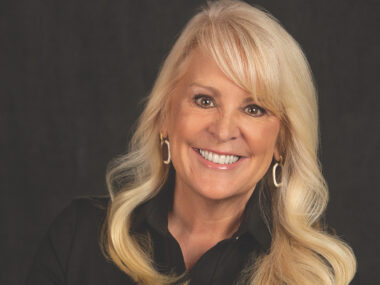 A headshot features a woman with blond hair smiling against a dark background.