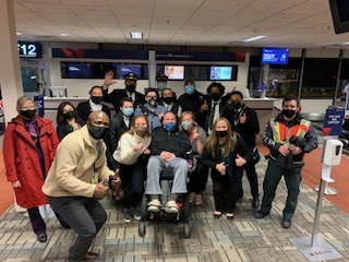 More than a dozen airline crew members father around a man in a wheelchair in front of a gate at the airport. They're all wearing face masks, and many are giving a double thumbs-up.