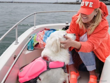 A woman wearing an orange jacket, baseball cap, and sneakers sits at the front of a boat with her dog, who's wearing a pink life jacket.