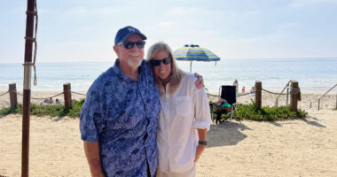 A man and woman pose for a photo under an umbrella in front of the beach.