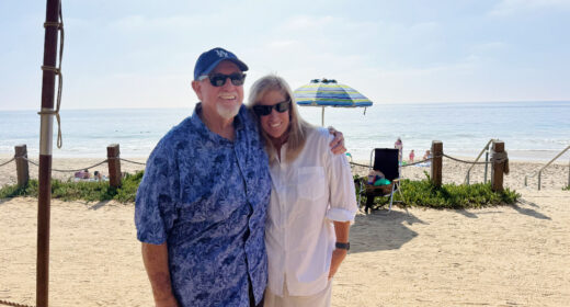 A man and woman pose for a photo under an umbrella in front of the beach.