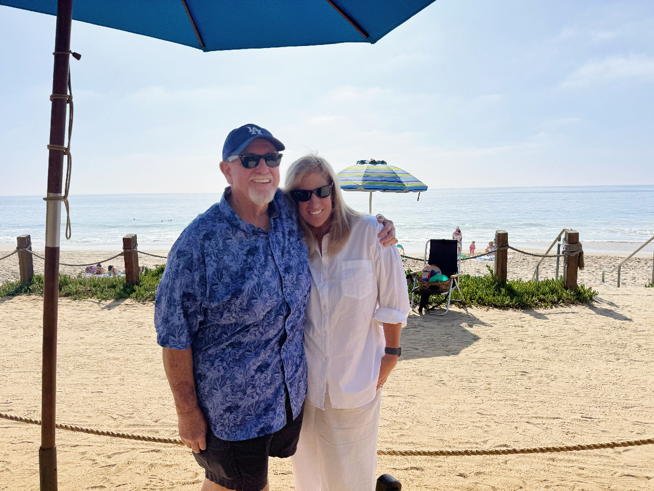 A man and woman pose for a photo under an umbrella in front of the beach.