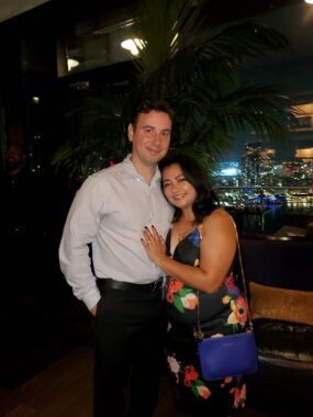 A man and a woman in dress clothes smile and embrace each other while standing in front of a bar. 