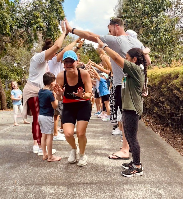 A woman in a baseball cap is seen coming out of the end of a human tunnel made by people clasping their hands together overhead.