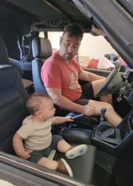A father and a toddler sit in a car. The father is in the driver's seat and looks down with a funny look at his son, who's sitting in the passenger seat and grabbing the parking break with his left hand. The photo shows only the interior of the car. 