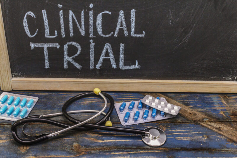 A stethoscope and oral medication packets are seen in front of a blackboard with the words "clinical trial" written on it in all capital letters.