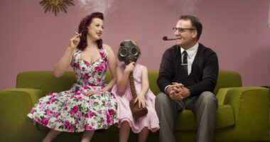 A child wearing a gas mask sites between a woman and man in 1950s attire who are smoking a cigarette and a pipe, respectively.