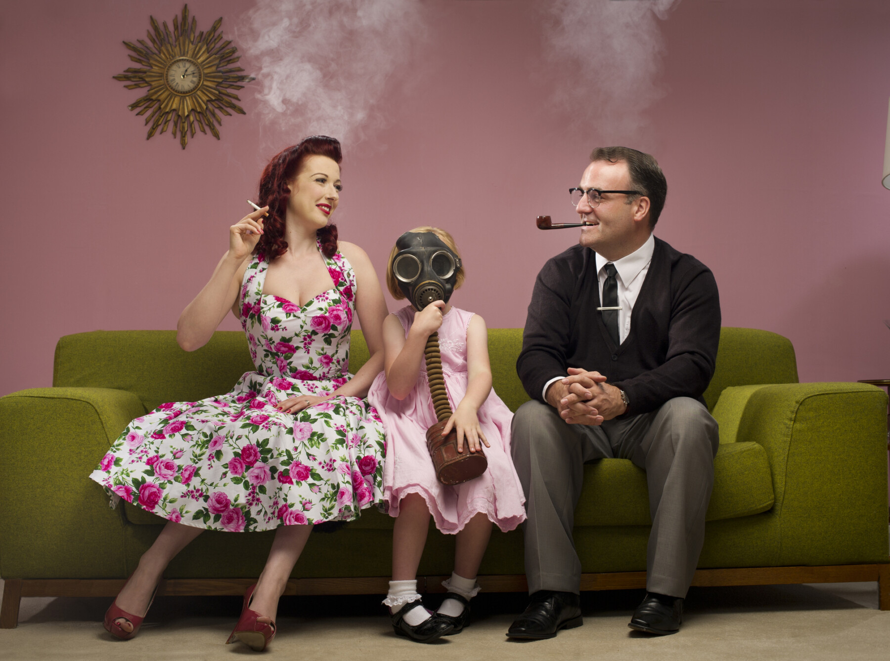 A child wearing a gas mask sites between a woman and man in 1950s attire who are smoking a cigarette and a pipe, respectively.