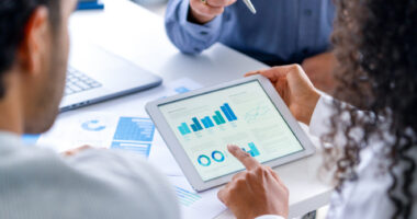 Three people look at data with graphs and charts shown on a tablet and papers with a laptop nearby.