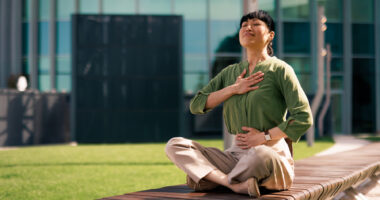 A woman is sitting on a bench practicing breathing exercises with her eyes closed and hands on her chest and stomach.