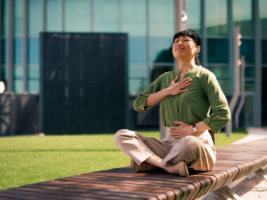 A woman is sitting on a bench practicing breathing exercises with her eyes closed and hands on her chest and stomach.