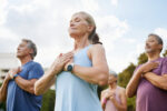 A group of people meditate outdoors, each with their hands on their heart.