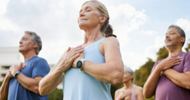 A photo of a group of people meditating peacefully outdoors in a park with hands on heart.
