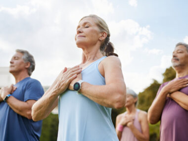 A photo of a group of people meditating peacefully outdoors in a park with hands on heart.