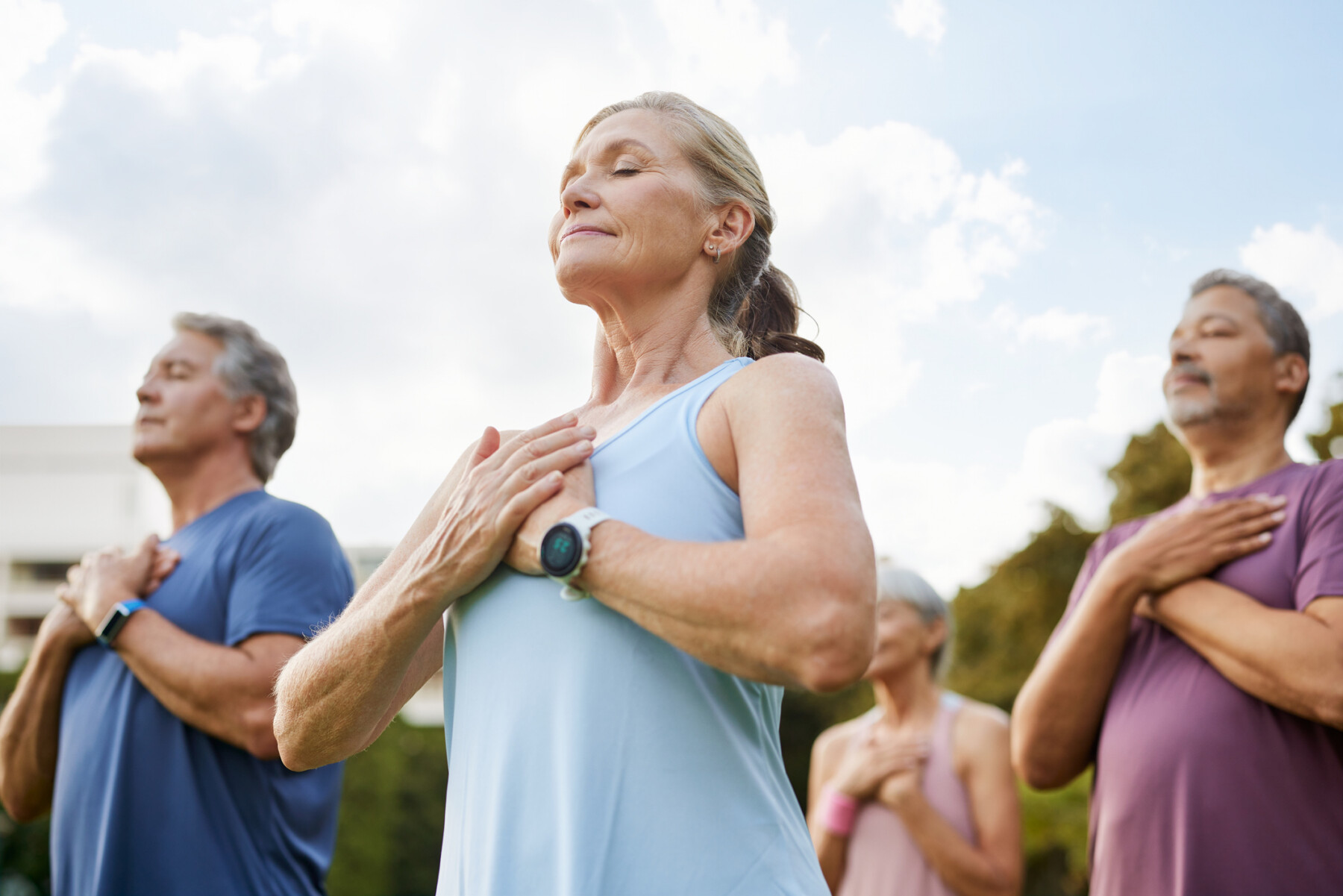 A photo of a group of people meditating peacefully outdoors in a park with hands on heart.