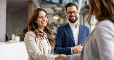 Two businesswomen shake hands as a businessman watches with his hands clasped.