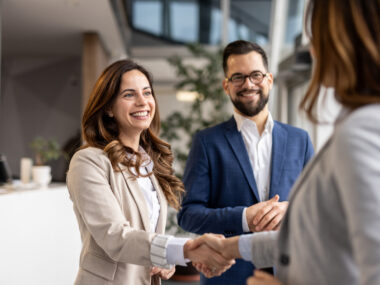 Two businesswomen shake hands as a businessman watches with his hands clasped.