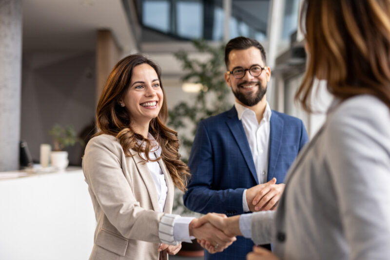 Two businesswomen shake hands as a businessman watches with his hands clasped.