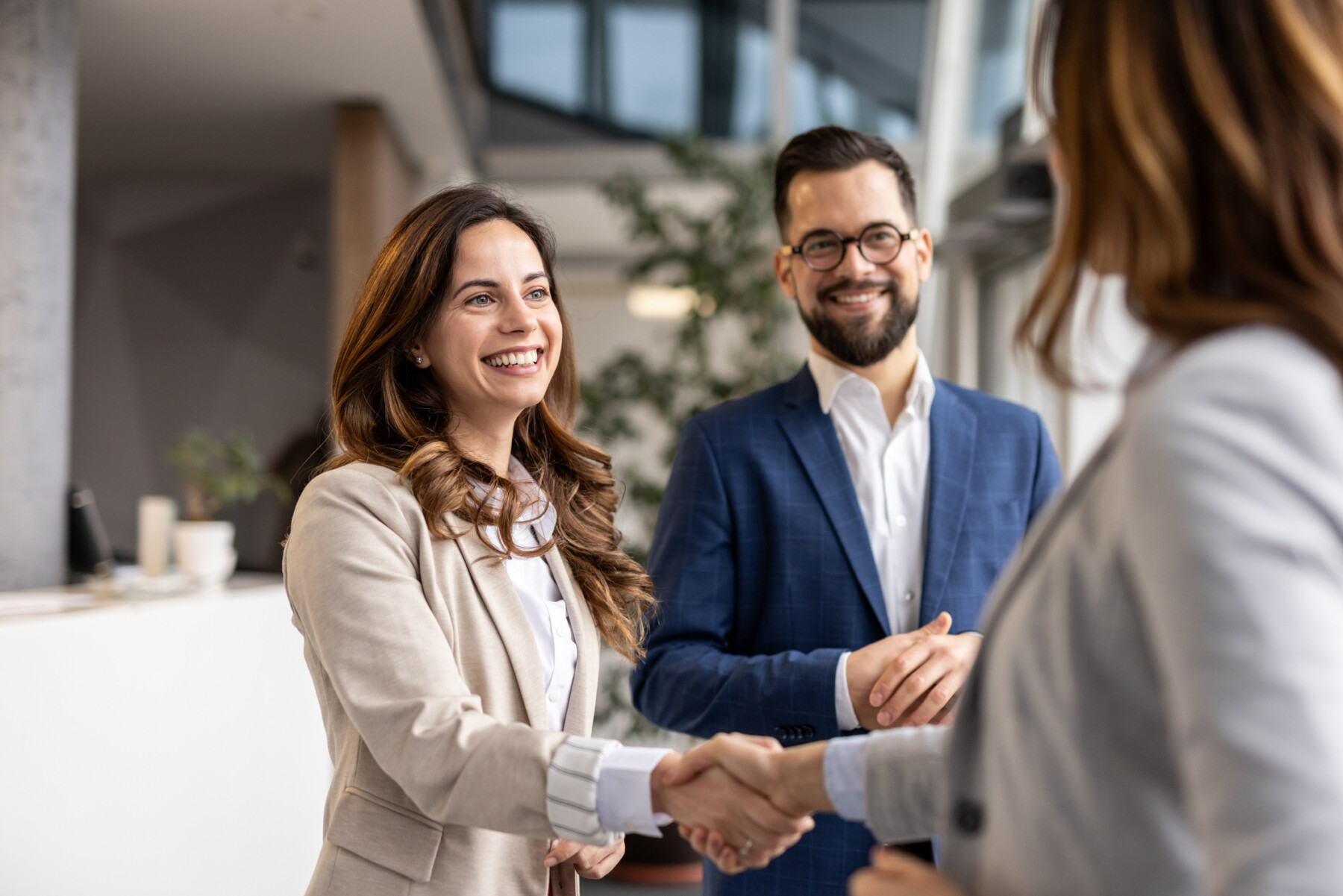 Two businesswomen shake hands as a businessman watches with his hands clasped.