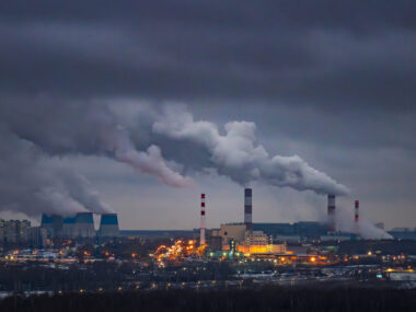 A panoramic view of an urban power plant, with multiple tall chimneys and cooling towers emitting thick plumes of smoke.