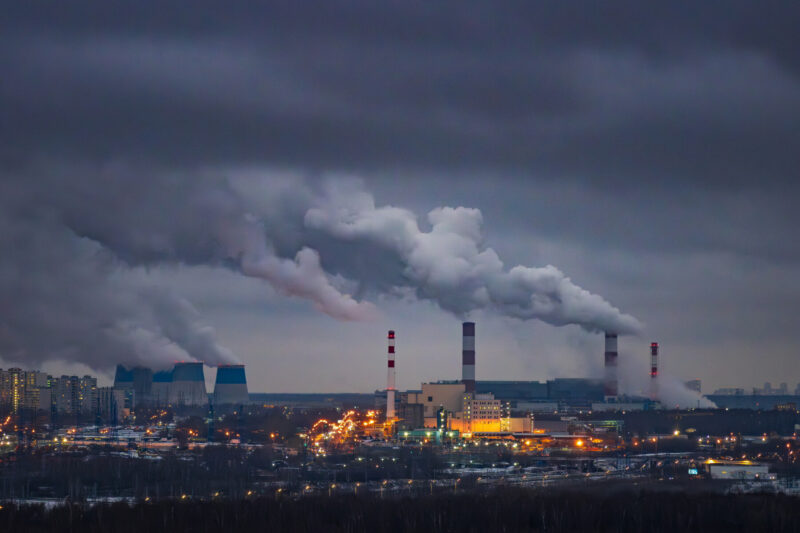A panoramic view of an urban power plant, with multiple tall chimneys and cooling towers emitting thick plumes of smoke.