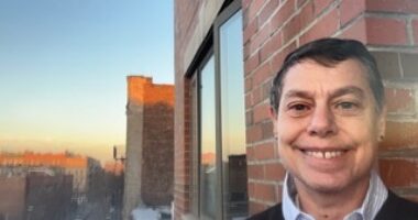 A woman stands on the deck of an apartment at what looks like sundown. She is smiling. A sweater indicates it may be brisk outside, and it appears she is in an urban area.