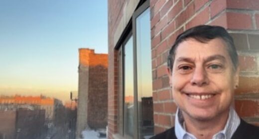 A woman stands on the deck of an apartment at what looks like sundown. She is smiling. A sweater indicates it may be brisk outside, and it appears she is in an urban area.