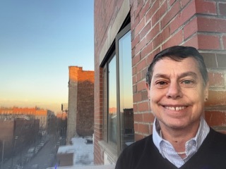 A woman stands on the deck of an apartment at what looks like sundown. She is smiling. A sweater indicates it may be brisk outside, and it appears she is in an urban area.