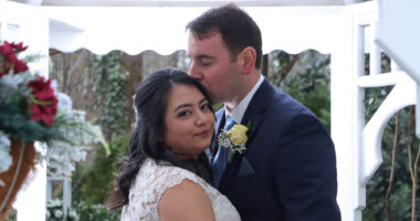 A man kisses his new wife on the head during their wedding. The bride is looking back at the camera over her right shoulder as the man looks down at her.