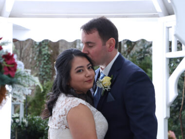 A man kisses his new wife on the head during their wedding. The bride is looking back at the camera over her right shoulder as the man looks down at her.