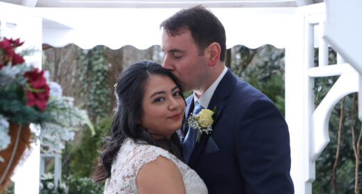 A man kisses his new wife on the head during their wedding. The bride is looking back at the camera over her right shoulder as the man looks down at her.