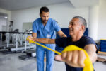 An image of a man taking part in physical therapy exercises using a stretch band with the assistance of his therapist.