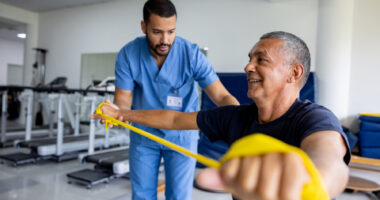 An image of a man taking part in physical therapy exercises using a stretch band with the assistance of his therapist.