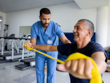 An image of a man taking part in physical therapy exercises using a stretch band with the assistance of his therapist.