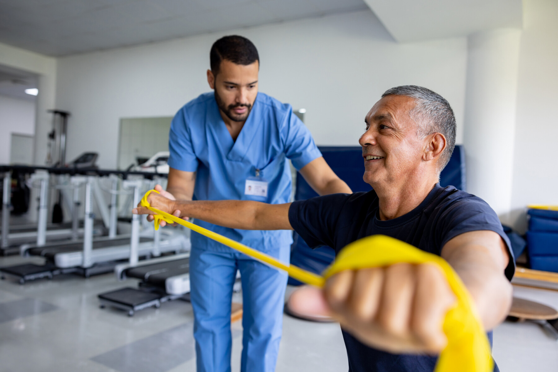 An image of a man taking part in physical therapy exercises using a stretch band with the assistance of his therapist.
