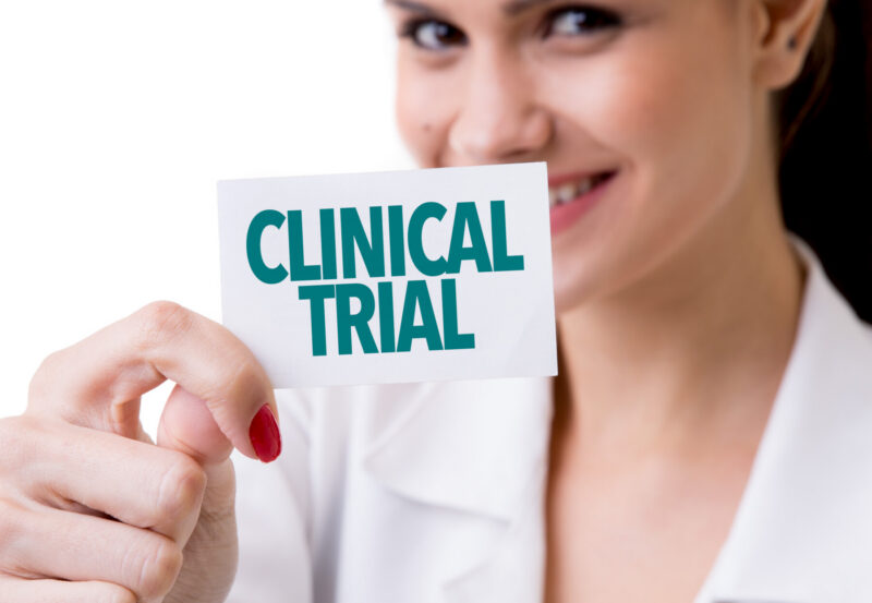 A smiling woman holds up a card with the words "clinical trial" printed on it in large green capital letters.