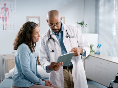 A doctor holds out a tablet for a patient to see while reviewing medical records in an exam room.
