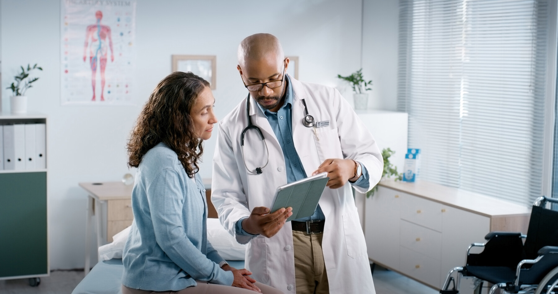 A doctor holds out a tablet for a patient to see while reviewing medical records in an exam room.