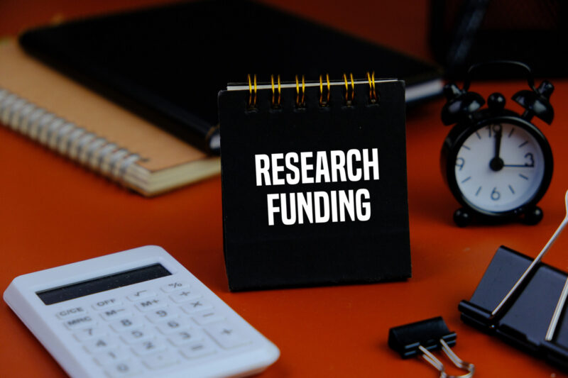 The words "RESEARCH FUNDING" are shown on a black notebook near a calculator.