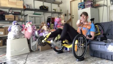 A man in cycling clothing and a worn baseball cap pops a wheelie in his recumbent trike in his garage while smiling. A gaggle of large, colorful stuffed animals sits to his right.