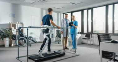 A patient wearing a robotic exoskeleton walks on a treadmill during physical therapy while being monitored by a pair of medical professionals. (Photo by iStock)