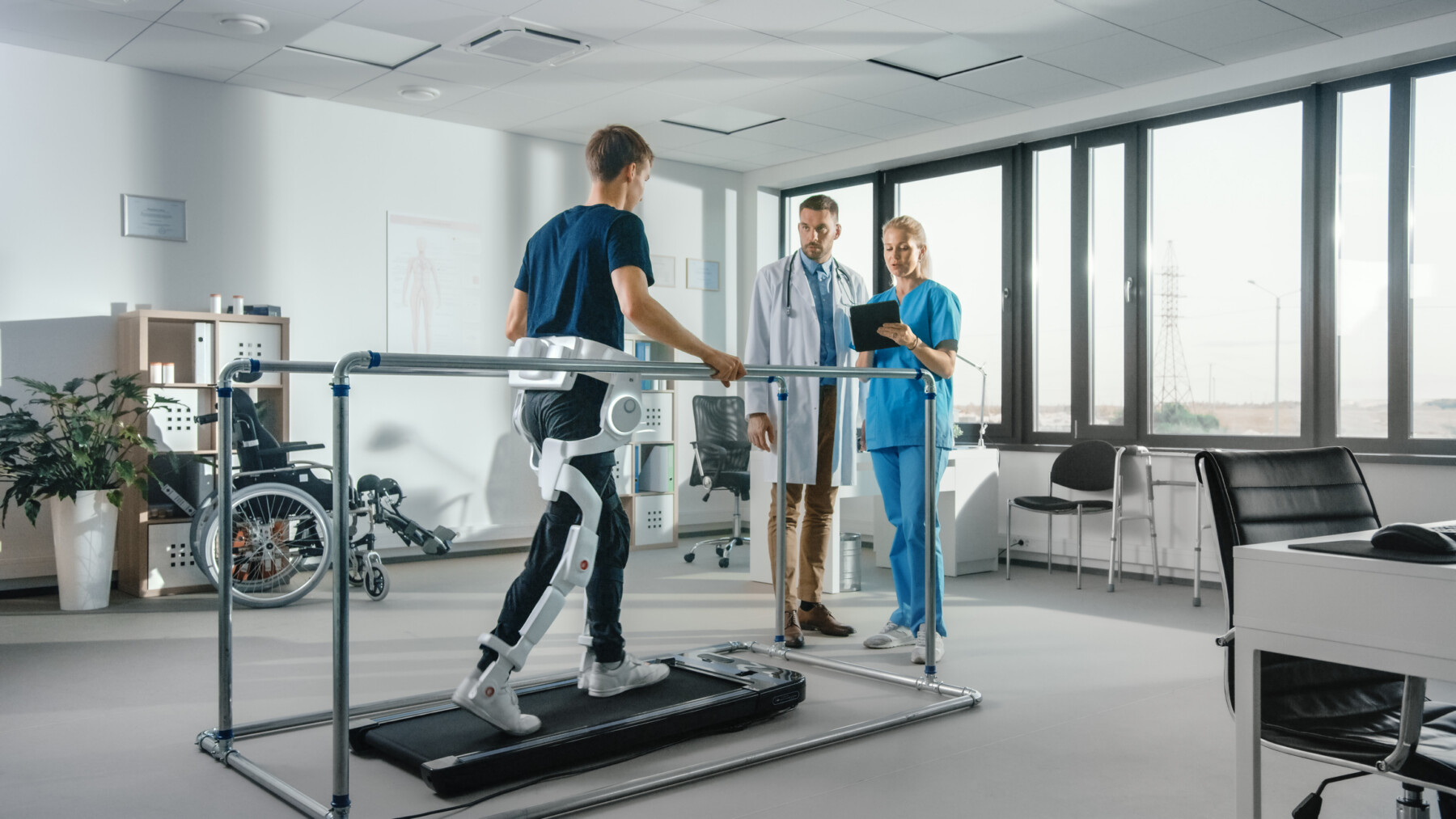 A patient wearing a robotic exoskeleton walks on a treadmill during physical therapy while being monitored by a pair of medical professionals. (Photo by iStock)