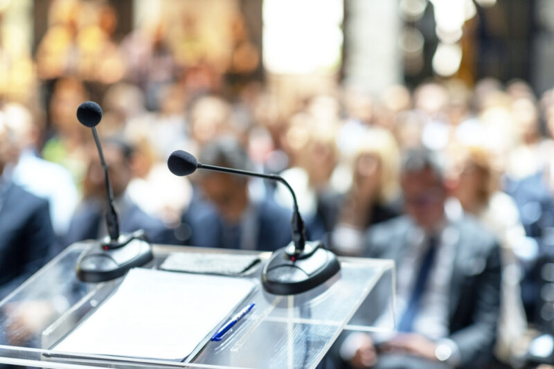 A microphone and some papers sit on a podium in front of a crowd.