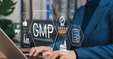 Icons for quality control and good manufacturing practices are seen above the hands of a person typing on a laptop keyboard.